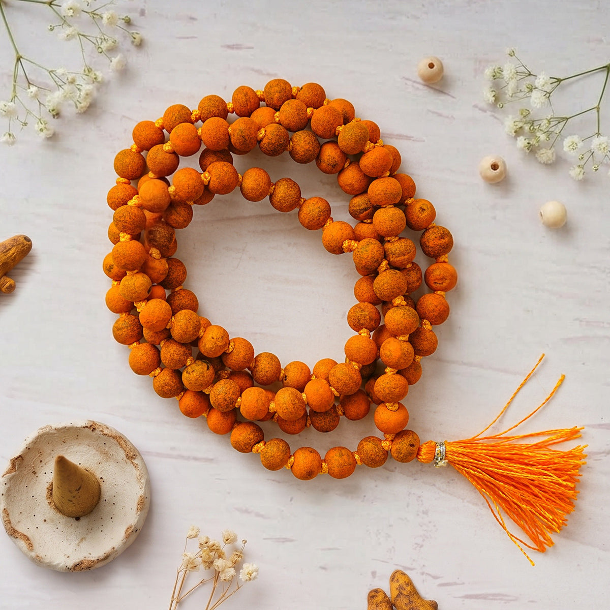 Natural Haldi Jaap Mala coiled on a white surface with an orange tassel, dried flowers and a dhoop cone