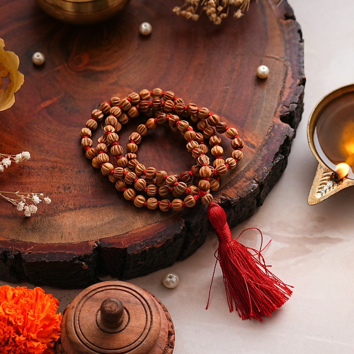 Chandan Carved Bead Mala coiled on a wooden slab beside a brass diya and pearls