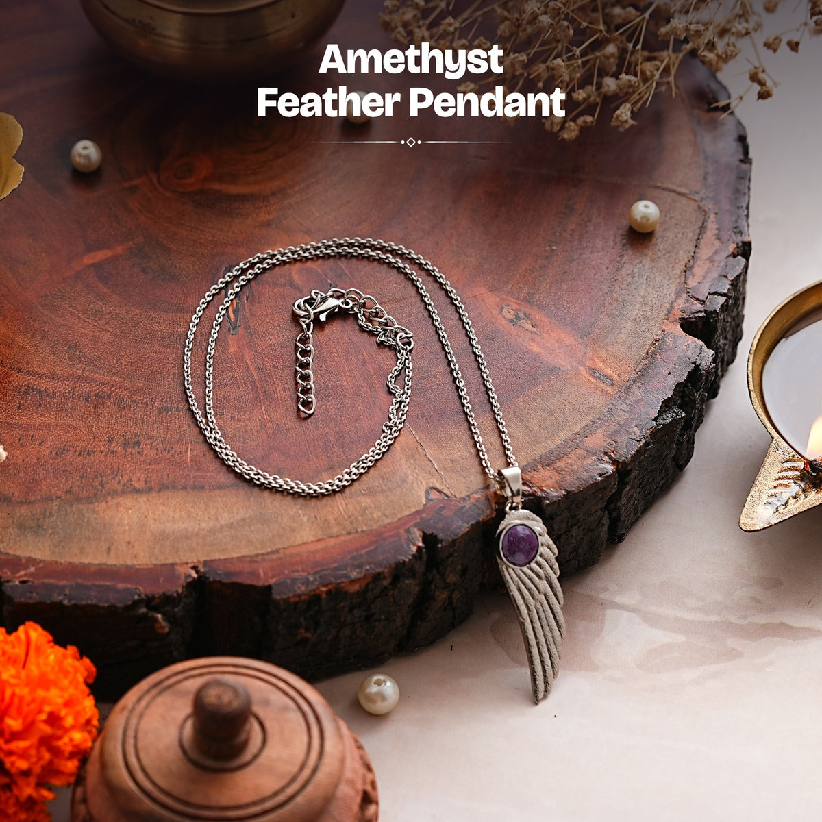 Amethyst feather pendant and chain on a wooden slab beside a lit diya and marigolds