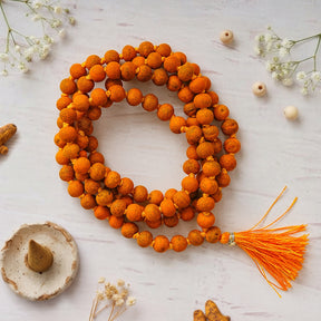 Natural Haldi Jaap Mala coiled on a white surface with an orange tassel, dried flowers and a dhoop cone