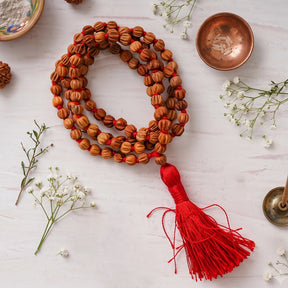 Chandan Carved Bead Mala coiled on a white surface with a red tassel, a copper bowl and baby's breath flowers