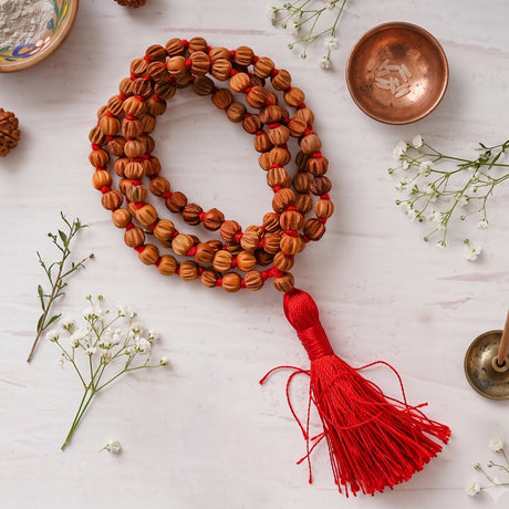 Chandan Carved Bead Mala coiled on a white surface with a red tassel, a copper bowl and baby's breath flowers