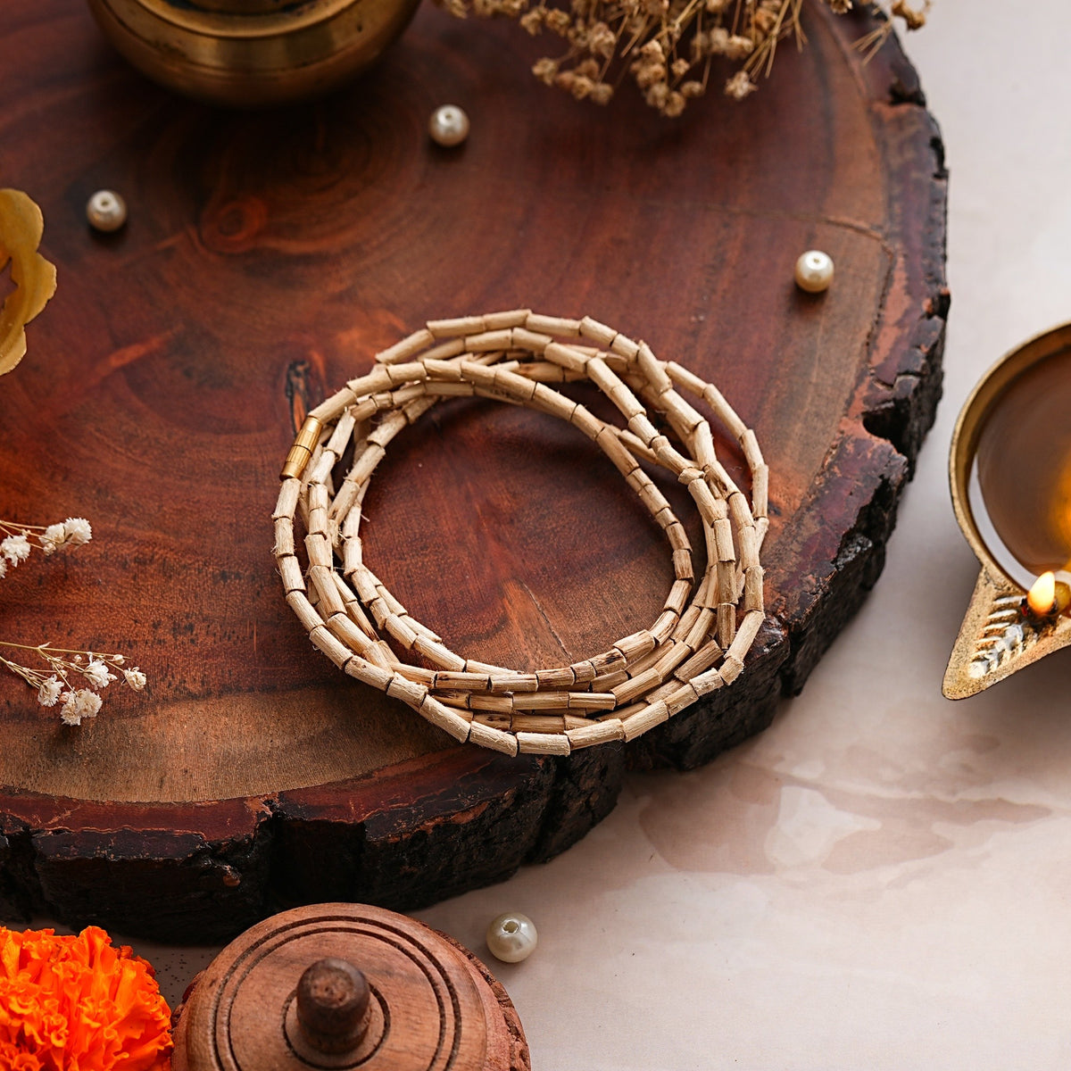 Natural Tulsi Kanthi Mala with elongated beads coiled on a wooden tray beside a diya and pearls